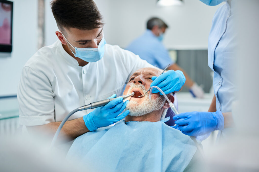 Young dentist polishing teeth of senior patient during dental procedure at clinic. Focus is on senior man.